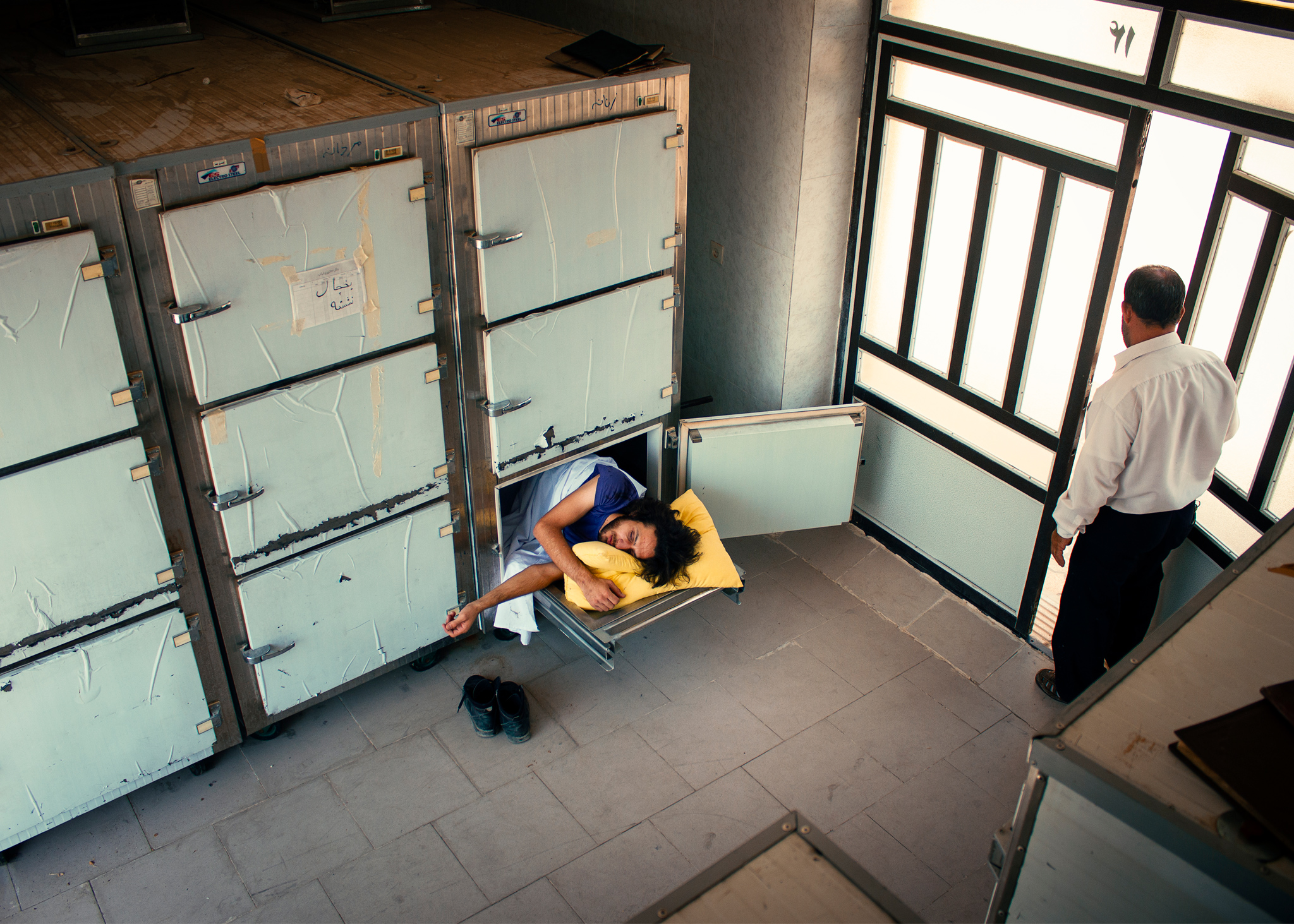 A man is lying inside an open morgue drawer, eyes closed, holding a yellow pillow. Another man stands by the door. Part of the conceptual photography series “Insomnia.”Insomnia #2: Inversion