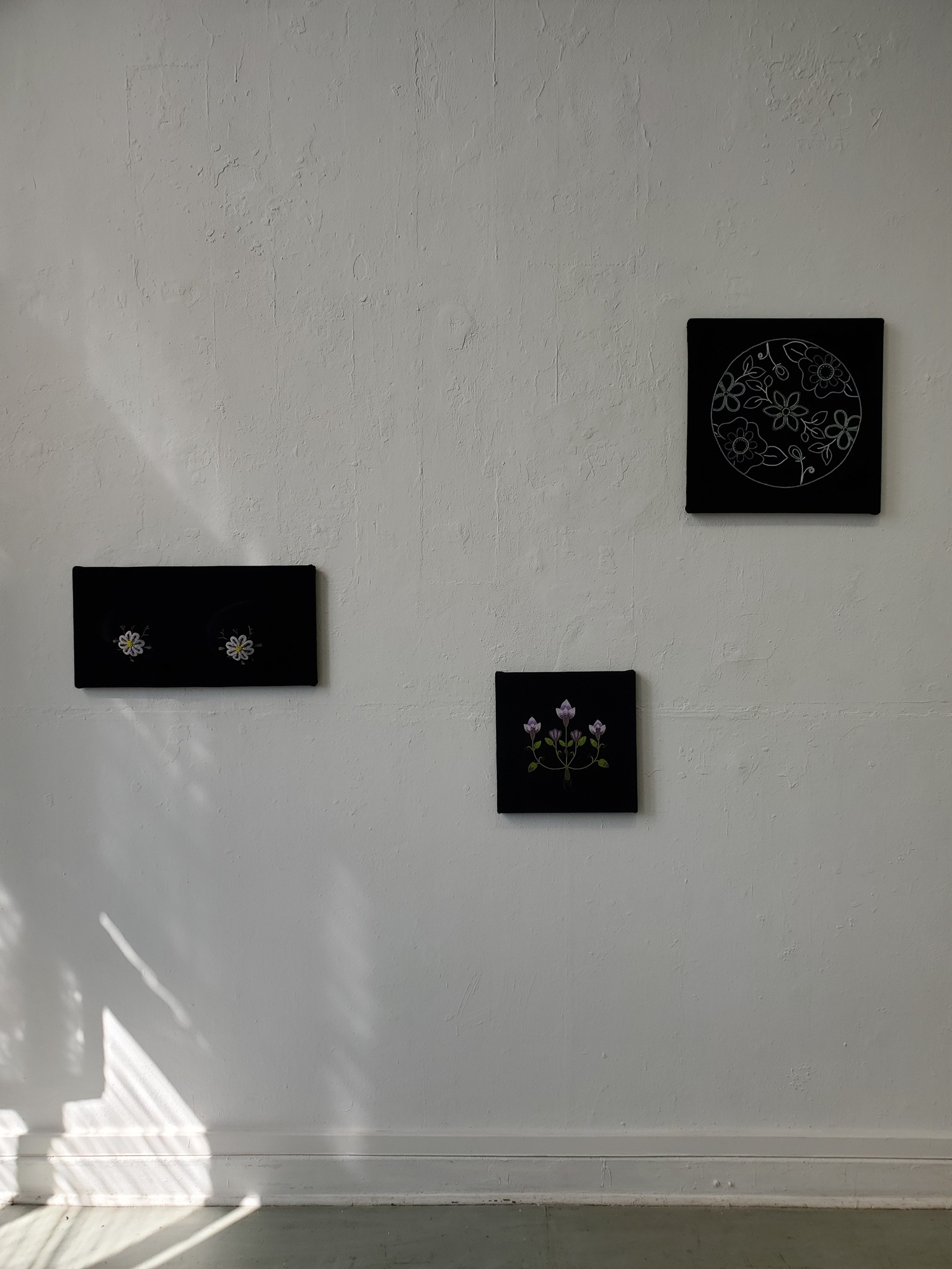 3 artworks are displayed across a while wall with sunlight shining below. furthest to the left a velvet bust attached to a rectangular backing, floral nipples made from glass beadwork and emboridery, next a smaller work on black wool depicting a 5 stem bundle of crocus flowers connected by a bulb and ebroidered roots. last a circular form places tor the right and above eye line. This piece is on black velvet ands depicts a moon shape with beaded outlines of flowers and foliage which become the craters of the moon.