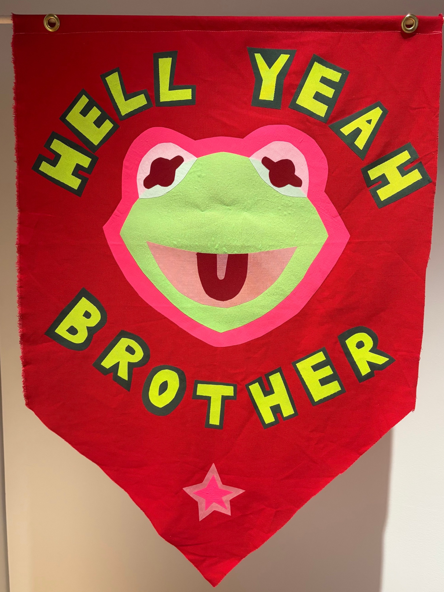 A Red flag with a fabric cut out of Kermit the frog on the front. Kermit is outlined in a bright pink fabric. Around Kermit's face is the phrase "HELL YEAH BROTHER" at the bottom of the tapered flag is a pink star. 
