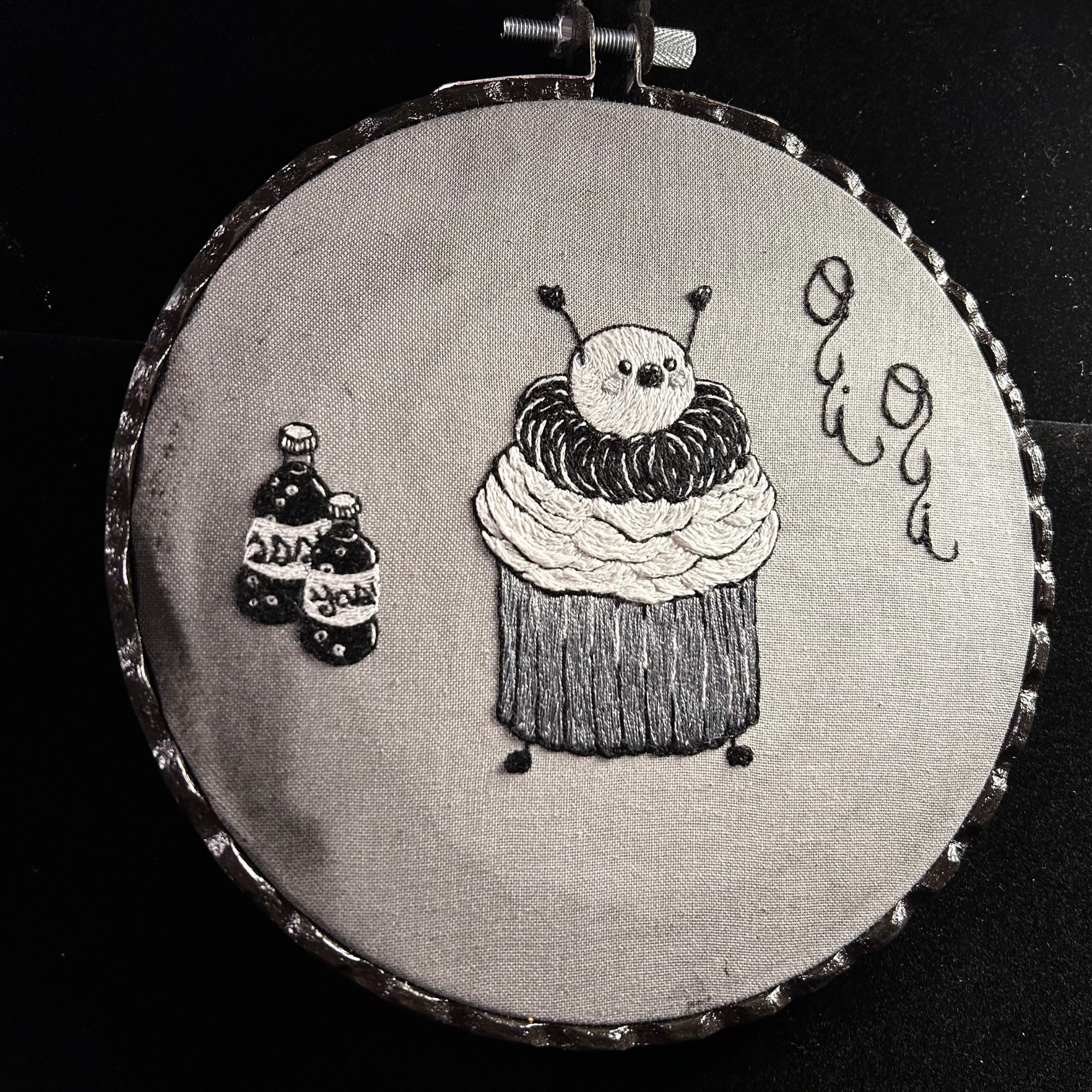 A black and white embroidery work that features a cupcake with little arms and legs. The cherry on the top of the cupcake acts as the head. The cupcake has a look of shock with it's arms thrown up. To the right of the cupcake sewing in cursive font are the words "oui, oui". To the left of the cupcake are two old fashioned bottles of bubbly soda.  